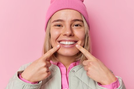 Eine junge blonde Frau mit pinker Mütze lächelt und zeigt mit ihren Fingern auf ihren Mund