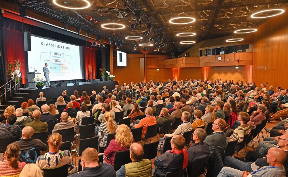 große Halle halbrunde Sitzreihen mit Publikum, Bühne mit Redner und Präsentation
