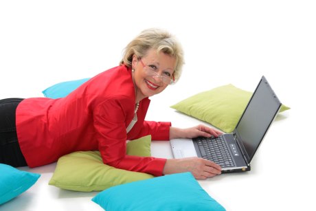 Frau um die 50 Jahre liegt lächeld auf dem Boden mit bunten Kissen und arbeitet am Laptop