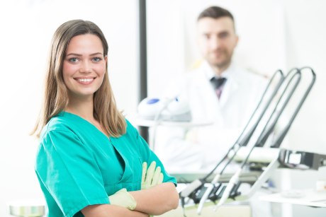 Ein Bild, das eine lächelnde junge Frau in grünem Zahnarzthelferin-Kittel und verschränkten Armen zeigt