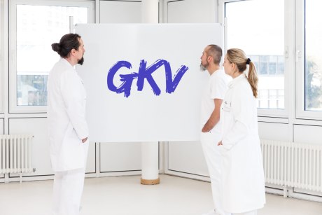 zwei Männer und eine Frau in Arztkitteln stehen vor einem Board mit den Buchstaben GKV in großen Lettern