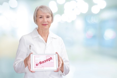 Ein Bild, das eine weißhaarige Frau in einem Weißen Kittel vor einem diffus hellen Hintergund zeigt. In der Hand hält sie ein Tablet mit einem Roten Schriftzugfür e-Rezept.