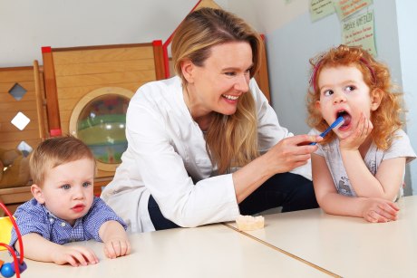 Ein Bild, das einen Kindergarteninnenraum mit Spielgeräten im Hintergrund zeigt. Im Vordergrund sitzt ein ins Nichtsstarrenden Dreijähriher im blaukarierten Hemd. Neben ihm putzt eine Frau im weißen Kittel einem vierjährigem Mädchen die Zähne.