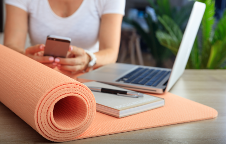 Yogamatte im Vordergrund mit Arbeitsbuch, Notebook und Frau am Handy dahinter