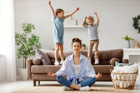 Mutter in Yogahaltung sitzt auf dem Boden, zwei Kinder toben hinter ihr auf einem Sofa