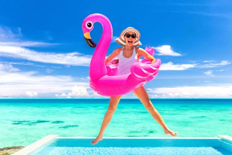 Frau in blau-weiß-gestreiftem Badeanzug mit Sonnenbrille und Sonnenhut und einem Flamingoschwimmreifen um die Taille springt vor einem Meer in die Luft