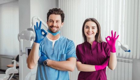 Mann und Frau in Arztkleidung in einer Zahnarztpraxis, die mit jeweils einer behandschuhten Hand das OK-Zeichen machen