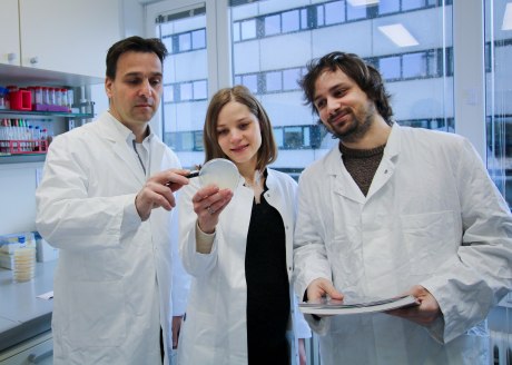 zwei Männer und eine Frau in weißen Kitteln in einem Raum, der aussieht wie ein Labor