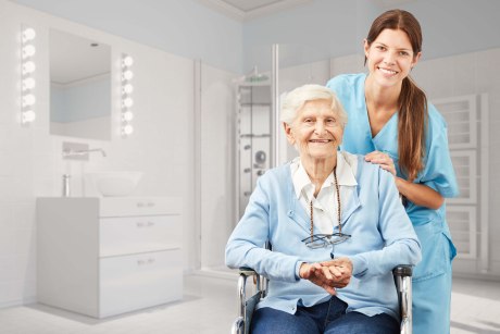 Eine junge Pflegekraft steht lächelnd hinter einer Seniorin, die im Rollstuhl sitzt
