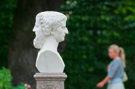 Ein Bild, das eine weiße Büste mit zwei Gesichtern im Profil zeigt. Im Hintergrund ist unscharf eine Frau, die durch einen Park geht.