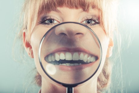 Ein Bild, das das Gesicht einer Frau zeigt, die sich eine Lupe vor den geöffneten Mund mit ihren Zähnen hält