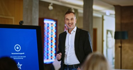 Ein Bild, das einen Mann mit weißem Hemd und blauem Jacket bei einer Präsentation in einem Innenraum zeigt.