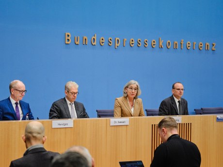 Vier Menschen sitzen hinter einer hölzernen Ballustrade vor einem blauen Fond, im Vordergrund sind ZUhören von hinten zu sehen.