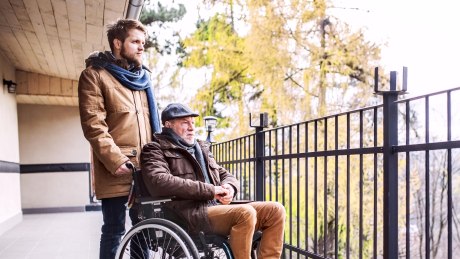 Pflegebedürftiger Mann im Rollstuhl schaut von einem Balkon in die Ferne, Pfeleger hinter ihm hält den Rollstuhl