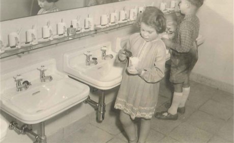 zwei Kinder mit Zahnpflege beschäftigt vor Waschbecken und einer langen Reihe von Zahnputzbechern in sw