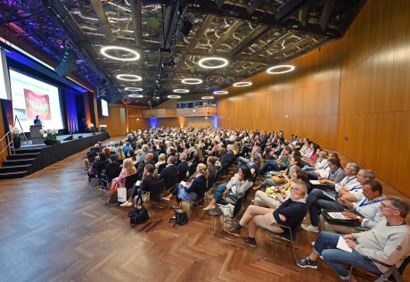 viele Menschen sitzen in einem großen Saal vor einer Bühne mit einer Präsentation