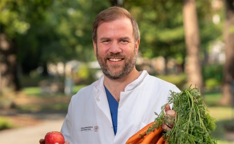 Prof. Dr. Johan Wölber in Artzkleidung mit einem Bund Karotten und einem Apfel in der Hand