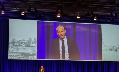 Dr. Christian Hammächer redend auf einer Bühne auf Leinwand projiziert