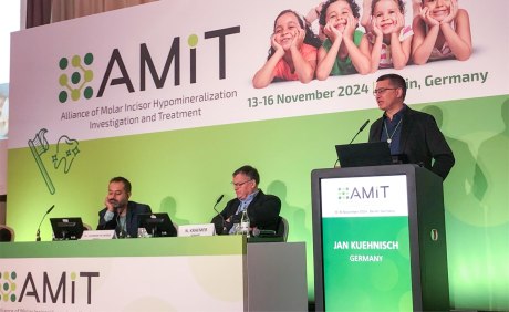 Jan Kuehnisch am Rednerpult auf dem Podium des AMIT-Kongresses mit 2 anderen Herren sitzend im Hintergrund