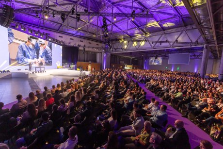 riesiger Saal mit sitzendem Publikum und Bühne mit Redner und Präsentation auf Leinwand
