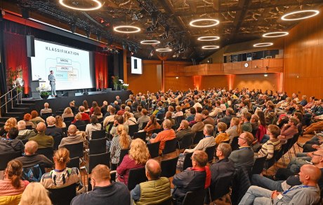 große Halle halbrunde Sitzreihen mit Publikum, Bühne mit Redner und Präsentation