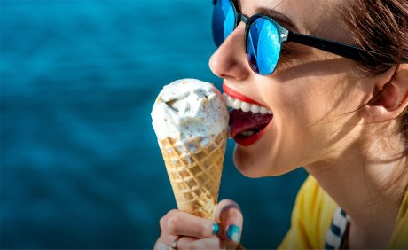 Frau mit Sonnenbrille vor blauem Hintergrund schleckt an einer Kugel Eis im Hörnchen