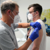 Älterer Mann gibt jüngerem Mann in einer Zahnarztpraxis eine Impfung in den Oberarm. Beide tragen Brille.