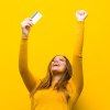 Junge Frau im gelben Pullover hält strahlend ihre Geldkarte in die Höhe
