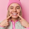 Eine junge blonde Frau mit pinker Mütze lächelt und zeigt mit ihren Fingern auf ihren Mund