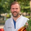 Prof. Dr. Johan Wölber in Artzkleidung mit einem Bund Karotten und einem Apfel in der Hand