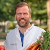 Prof. Dr. Johan Wölber in Artzkleidung mit einem Bund Karotten und einem Apfel in der Hand