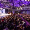 riesiger Saal mit sitzendem Publikum und Bühne mit Redner und Präsentation auf Leinwand