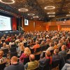 große Halle halbrunde Sitzreihen mit Publikum, Bühne mit Redner und Präsentation