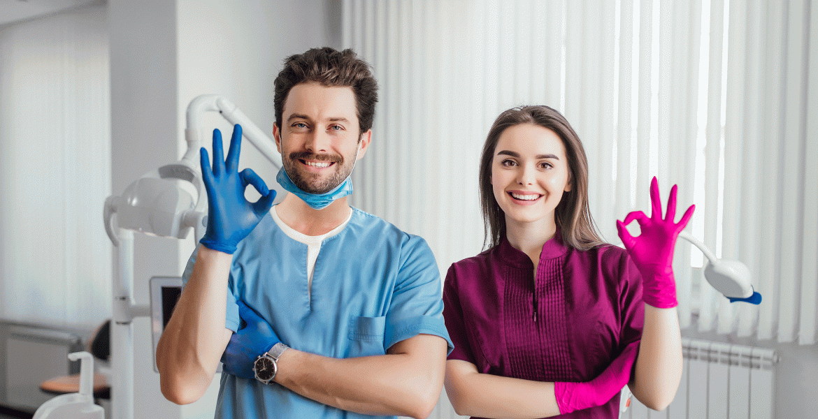 Mann und Frau in Arztkleidung in einer Zahnarztpraxis, die mit jeweils einer behandschuhten Hand das OK-Zeichen machen