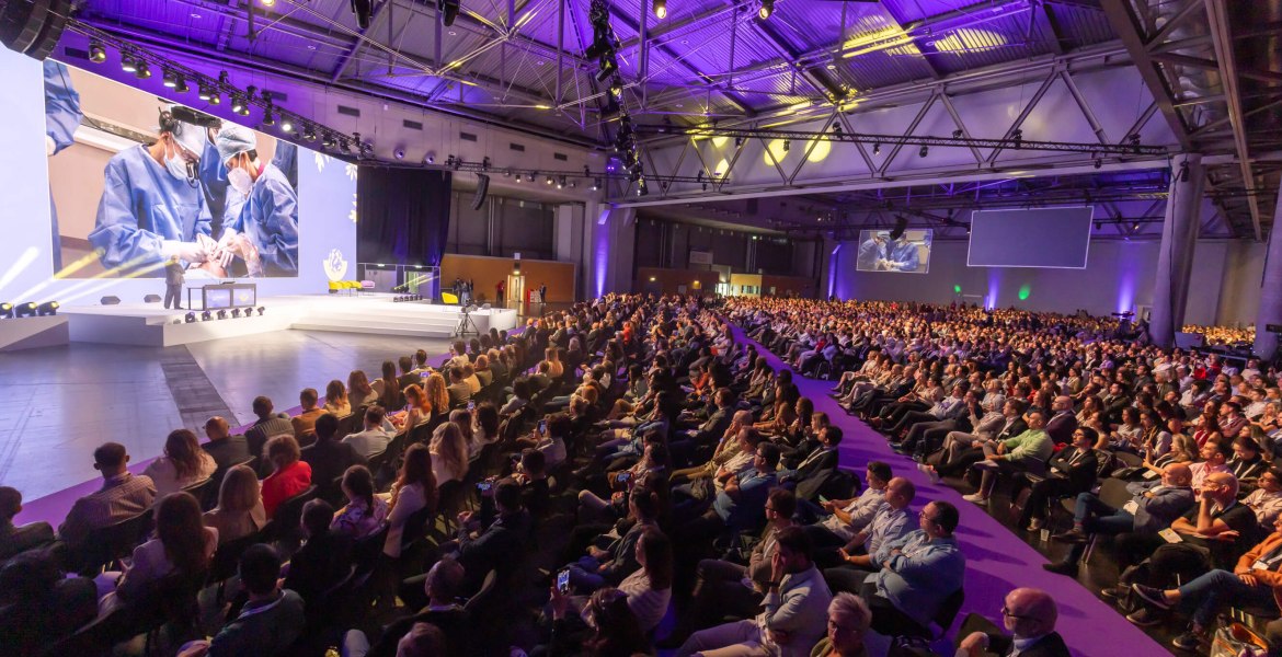 riesiger Saal mit sitzendem Publikum und Bühne mit Redner und Präsentation auf Leinwand