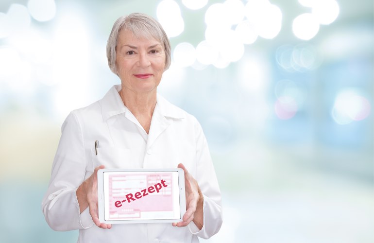 Ein Bild, das eine weißhaarige Frau in einem Weißen Kittel vor einem diffus hellen Hintergund zeigt. In der Hand hält sie ein Tablet mit einem Roten Schriftzugfür e-Rezept.