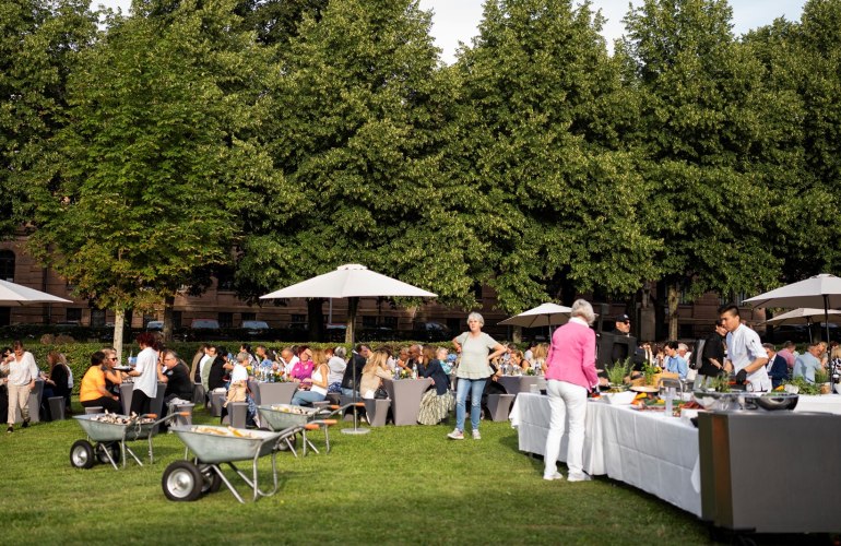 Wiese, viele Tische und Bänke mit Menschen, Buffet, Schubkarren mit Getränken