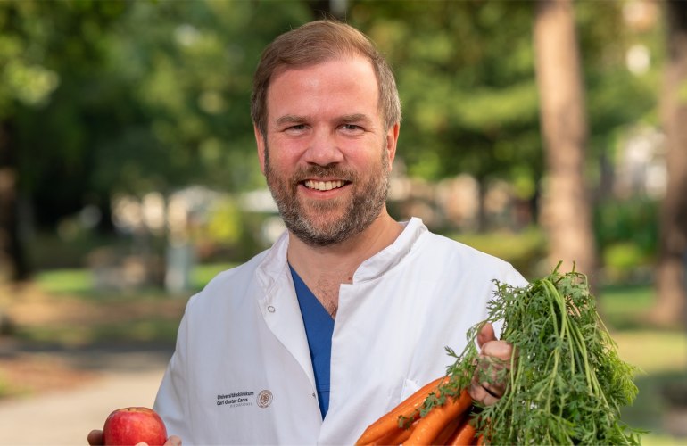 Prof. Dr. Johan Wölber in Artzkleidung mit einem Bund Karotten und einem Apfel in der Hand