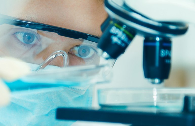 Mensch mit Schutzbrille und Atemschutz hält vermutlich eine Pinzette mit kreisförmigem Objekt unter Mikroskop