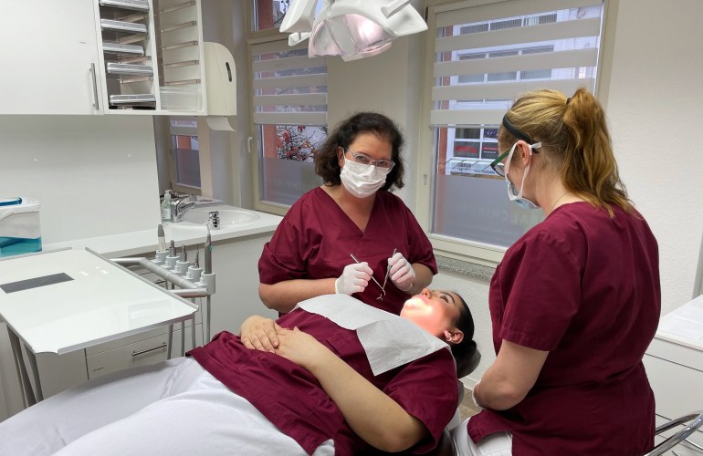 Frauen in medizinischer Kleidung und Maske beugen sich über Patientin auf Behandlungsstuhl