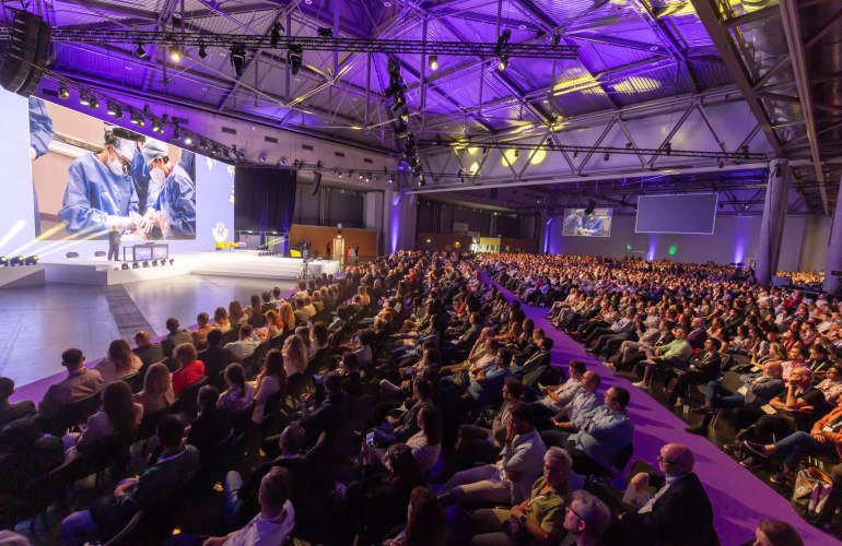 riesiger Saal mit sitzendem Publikum und Bühne mit Redner und Präsentation auf Leinwand